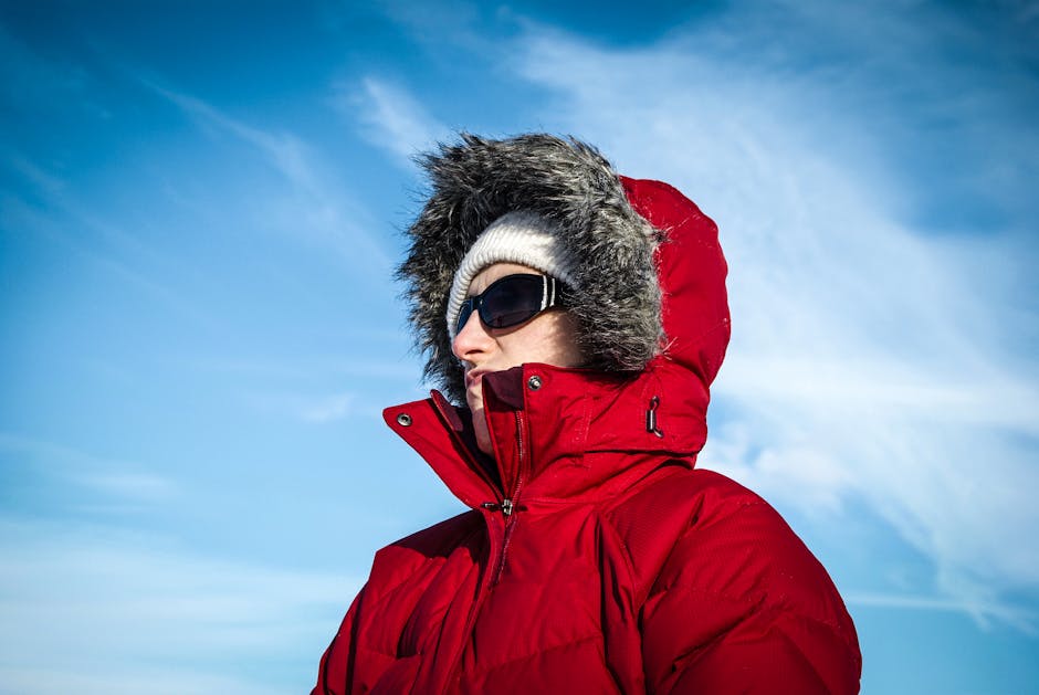 A person in a red winter coat and sunglasses stands against a clear blue sky.