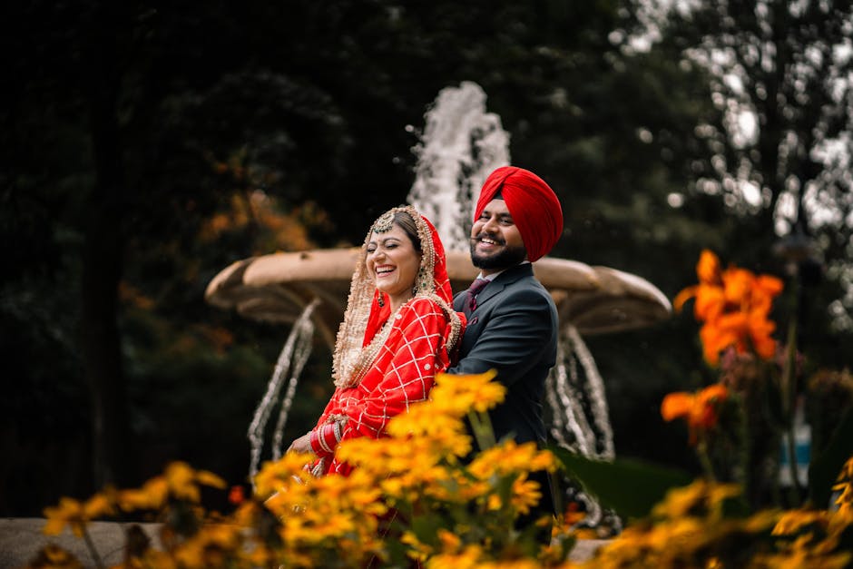 Sikh bride and groom in traditional attire smiling by a fountain surrounded by flowers in Brampton.