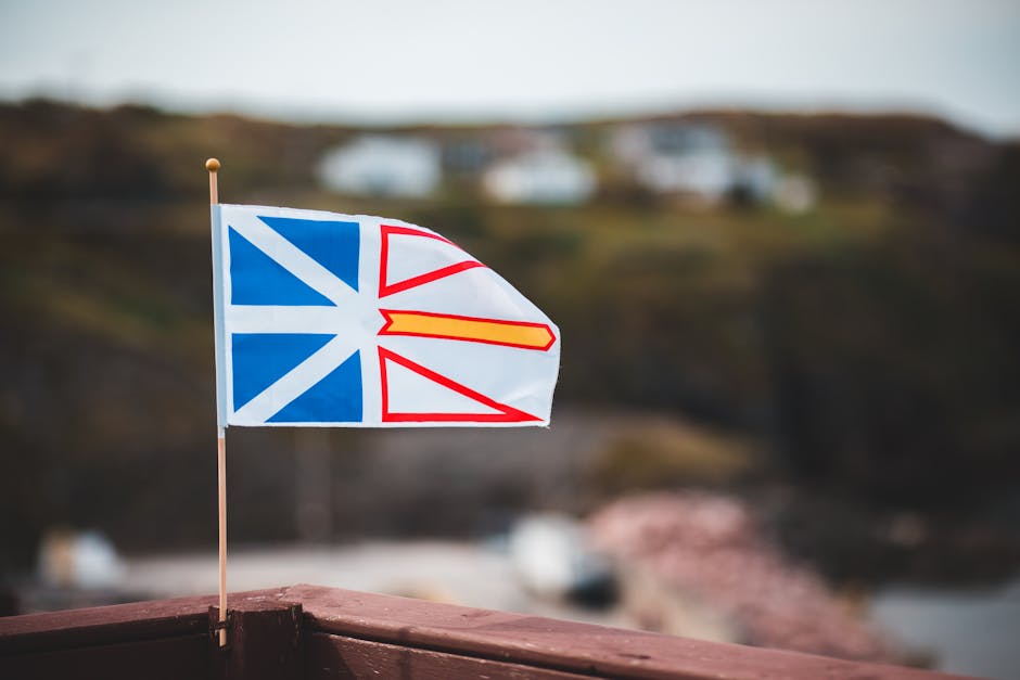 Waving national flag of Newfoundland and Labrador placed on wooden fence against blurred coastal settlement on hilly ter