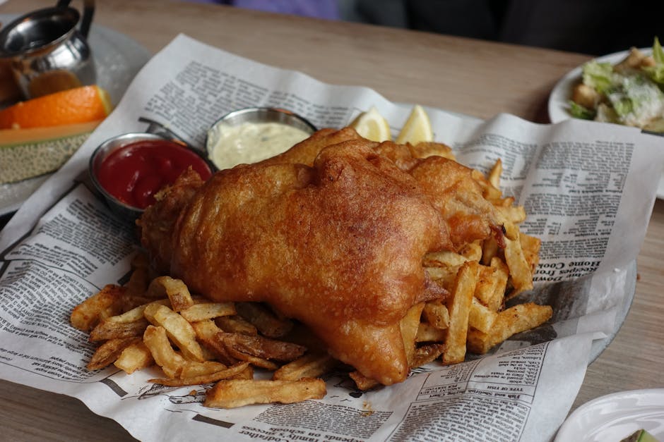 Delicious crispy fish and chips served with red and white dipping sauces on a newsprint lined platter.
